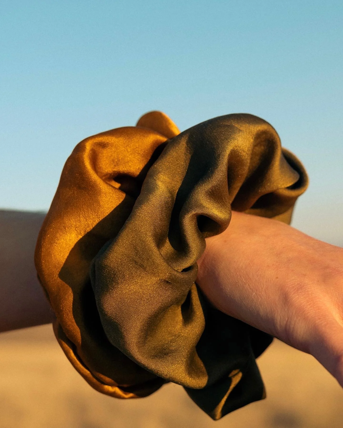 Close-up of a person's arm wearing a large, golden silk scrunchie against a blue sky background.