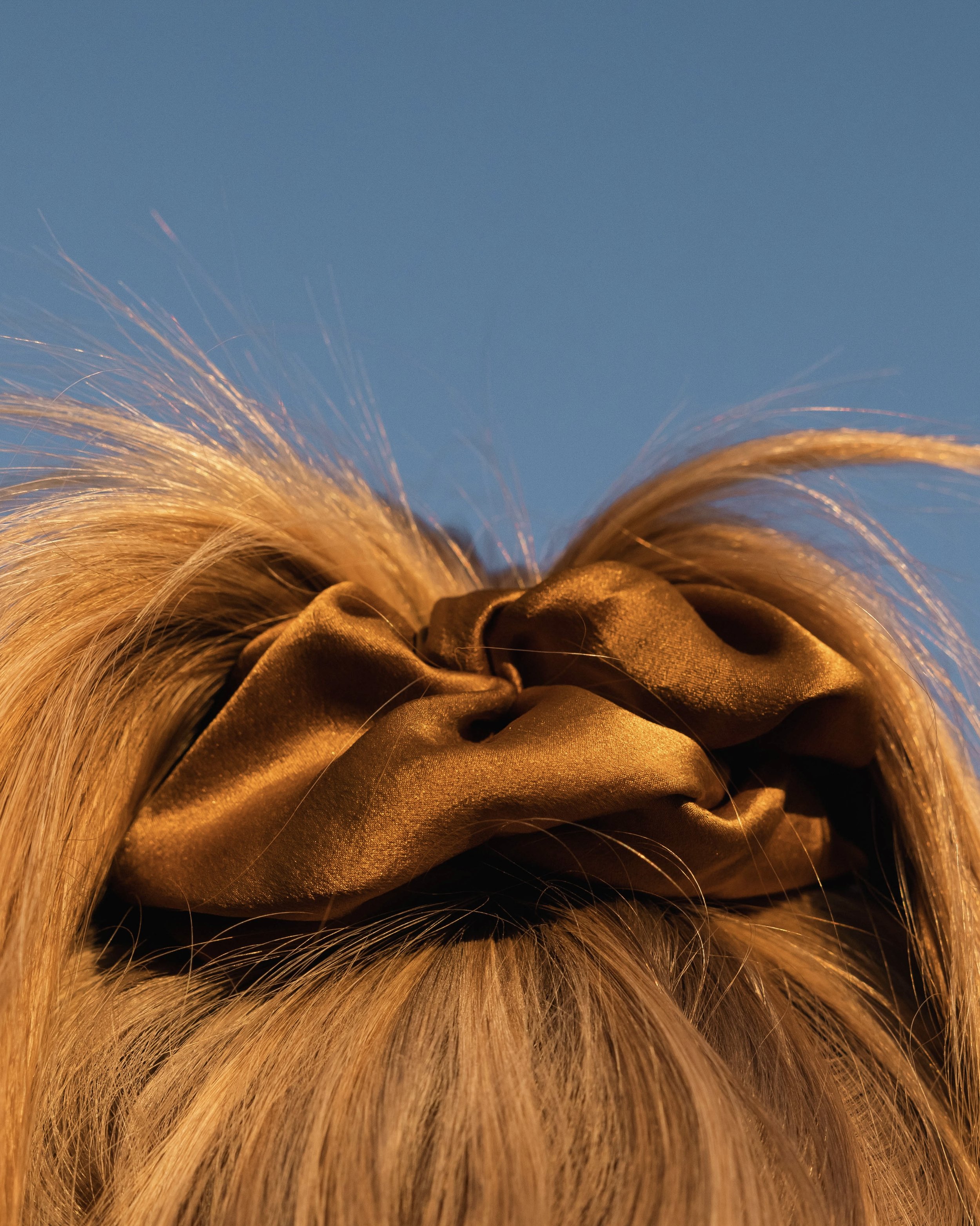 Close-up of a brown hair scrunchie holding blonde hair in a ponytail against a clear blue sky.