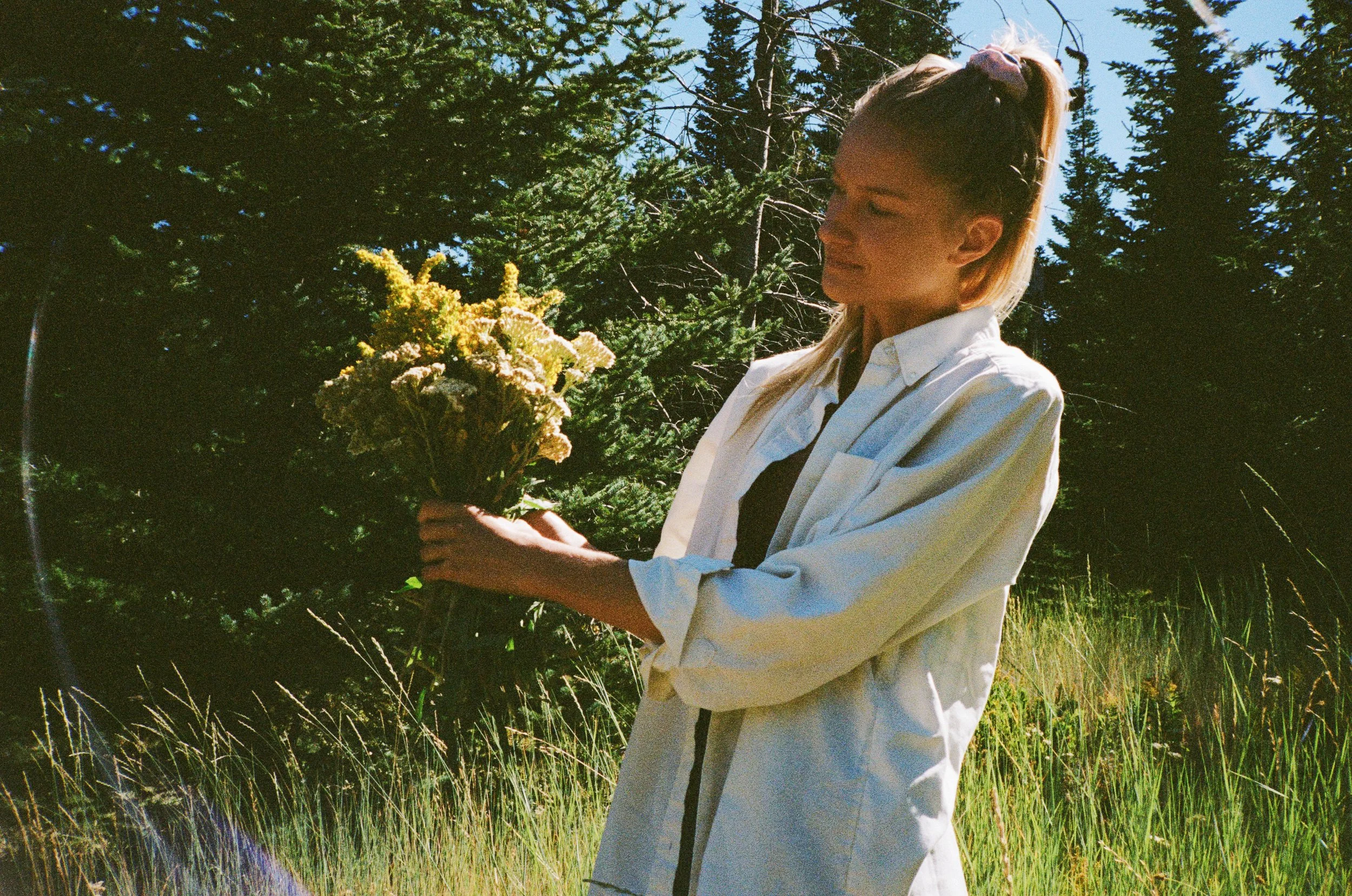 Woman in white shirt holding a bouquet of wildflowers in a grassy area with trees in the background.