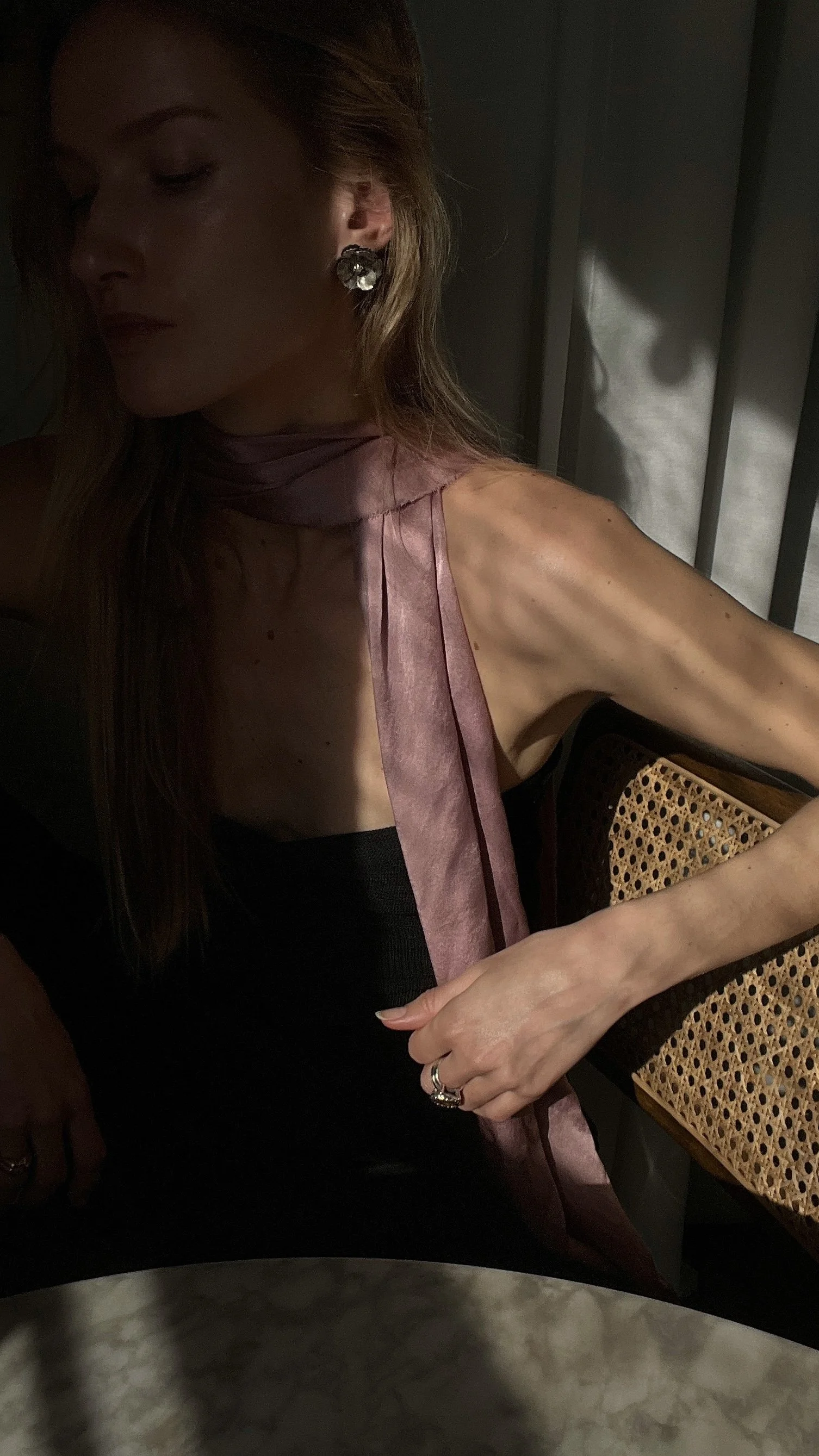 Woman seated in shadows, wearing a scarf and earrings, with a woven chair and marble table visible.