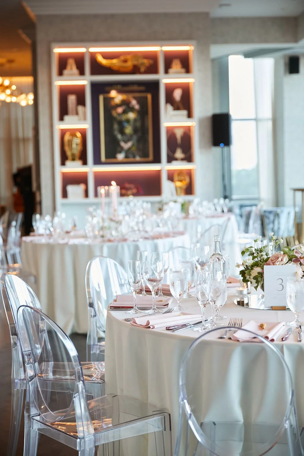 Elegant dining setup in a restaurant with round tables covered in white cloths, clear chairs, neatly arranged glassware, pink napkins, table numbers, and floral centerpieces. Background features illuminated shelves with decorative items.