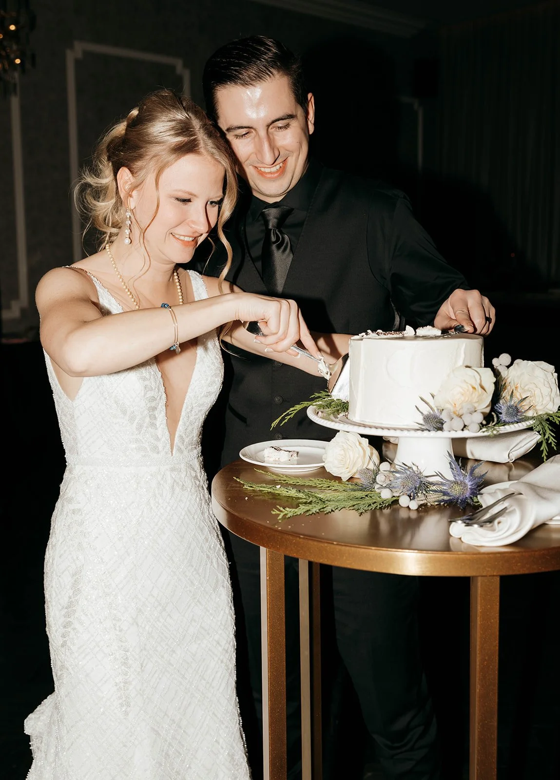 A bride and groom smiling as they cut a wedding cake together. The cake is decorated with white frosting and surrounded by flowers and greenery on a gold table.
