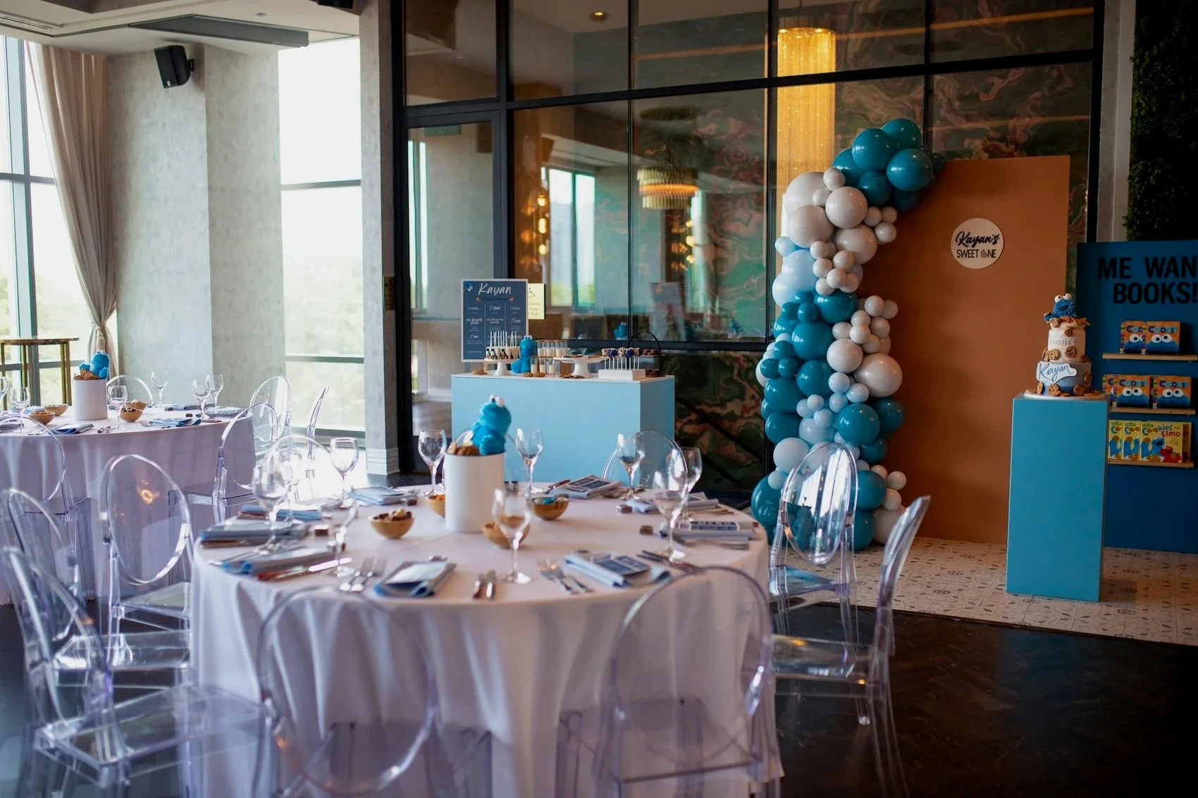 Party setup with round tables covered in white tablecloths, clear plastic chairs, and blue and white decorations. There are plates, silverware, glasses, and small bowls on the tables. A balloon arch made of blue and white balloons stands on a wall section, with a sign that reads 'Kayan's Sweet One'. A dessert table has themed treats and a cake decorated with blue and brown accents, featuring Cookie Monster. In the background, there are large windows and a cozy, modern interior.