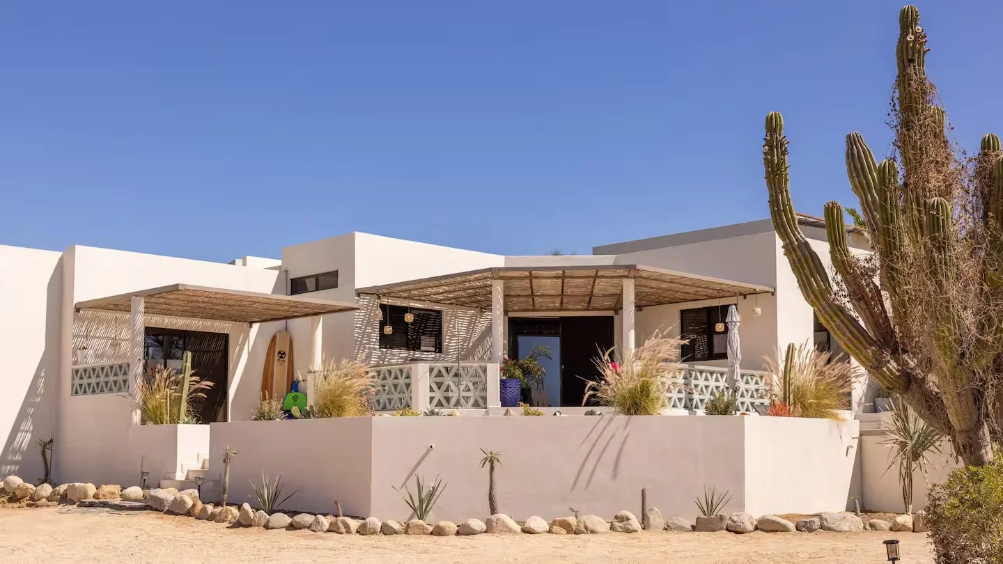 A modern white house with a patio, surrounded by desert plants and cactus, under a clear blue sky.