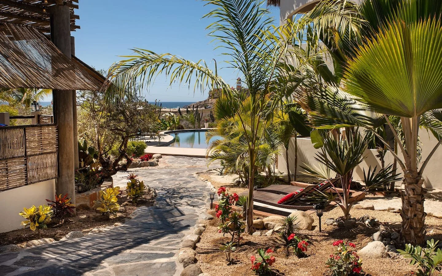 A tropical garden with palm trees, flowering plants, and a stone pathway leading to a backyard pool with ocean view.