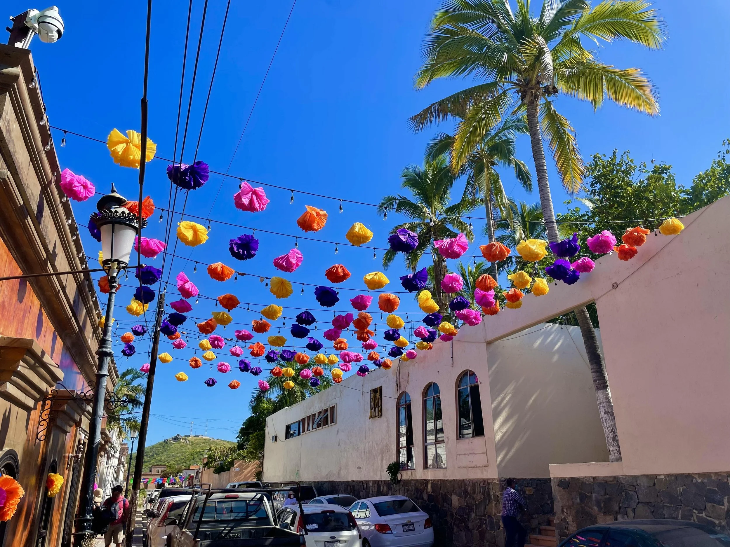 Colorful paper lanterns hanging above a busy street in a sunny area with palm trees and buildings.