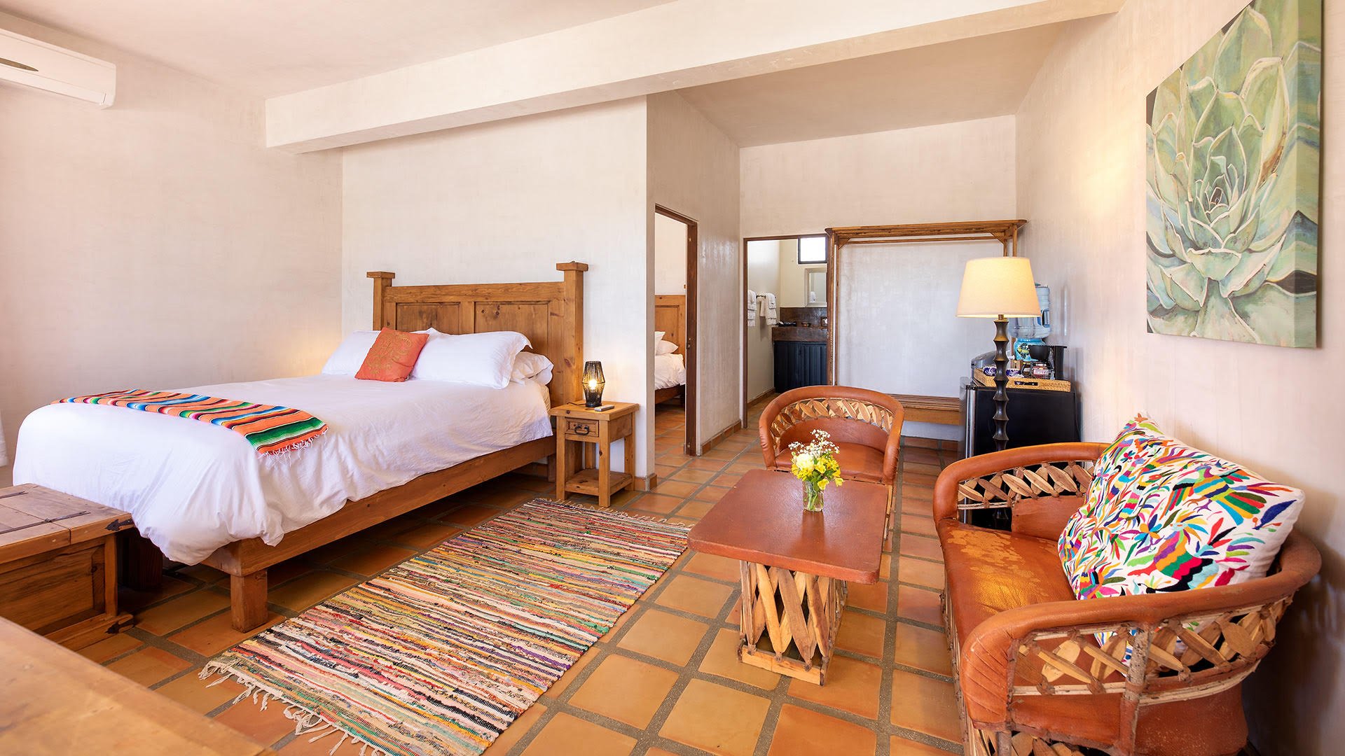 A cozy bedroom with a wooden bed, colorful textiles, a small sitting area, and a kitchenette in the background.