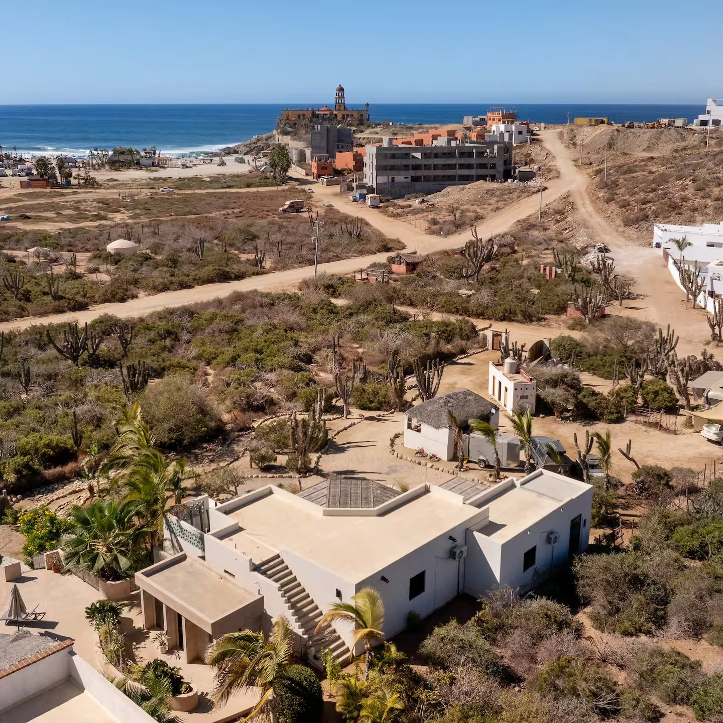 Aerial view of a coastal area with white buildings, desert vegetation, dirt roads, and a view of the ocean with a lighthouse.