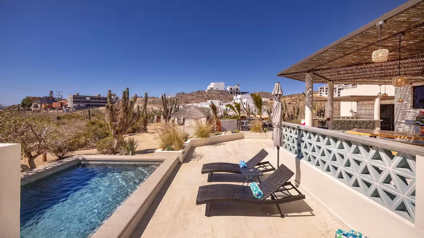 A sunny outdoor patio with two black lounge chairs with rolled-up towels, a small pool, and a shaded seating area with wooden accents, in a desert landscape with cacti and dry terrain in the background.