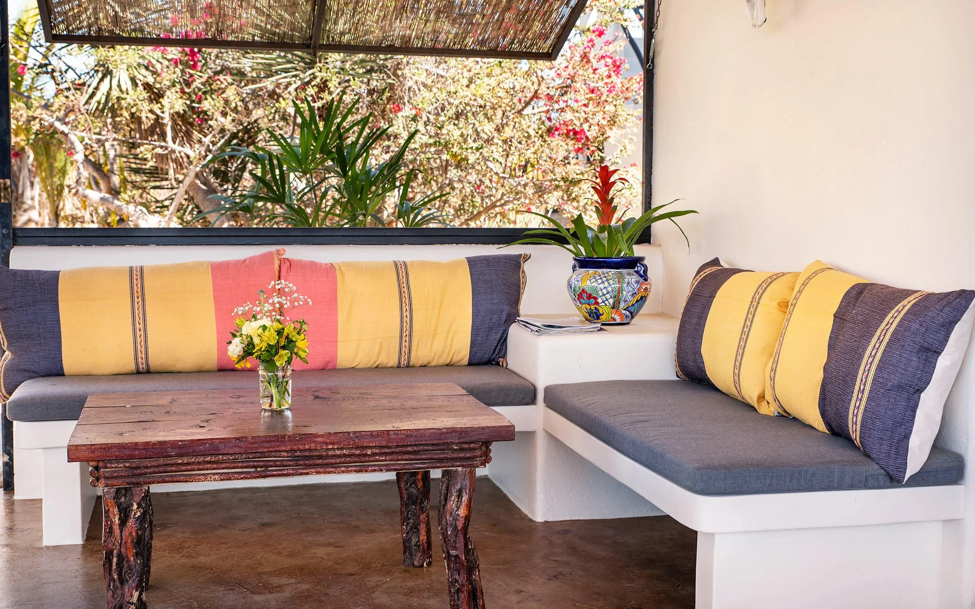Indoor seating area with a wooden table, colorful cushions, potted plants, and a view of trees outside through an open window.