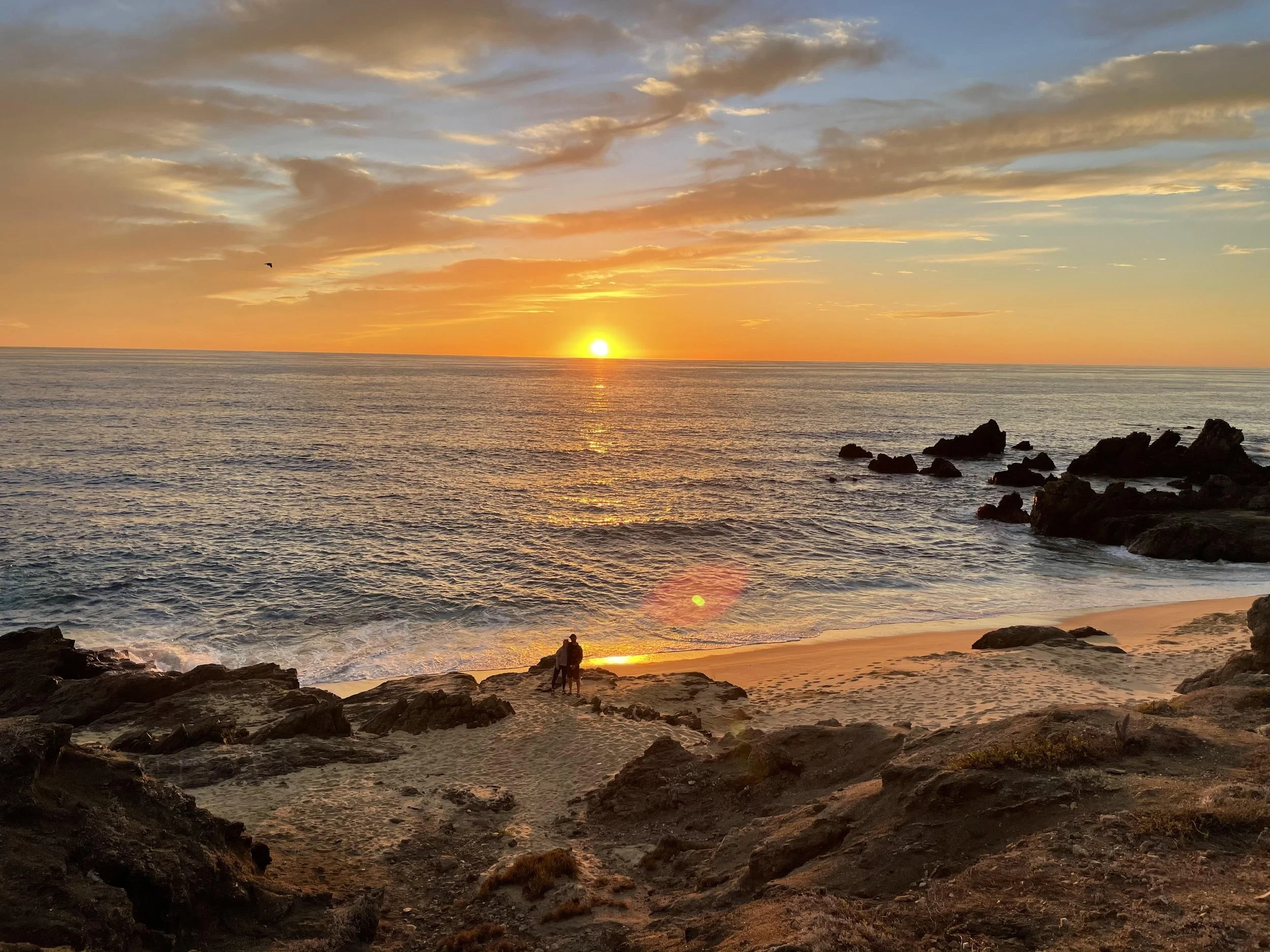A sunset scene over the ocean with a partly cloudy sky, rocks on the beach, and a couple standing on the rocks amidst the sunset glow.