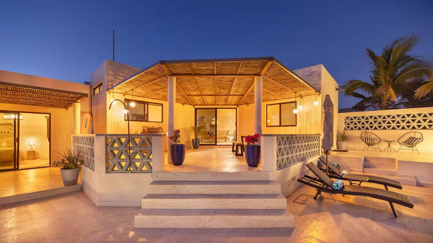 A modern outdoor terrace of a house at twilight, with steps leading up to a covered patio area, lounge chairs, potted plants, and palm trees in the background.