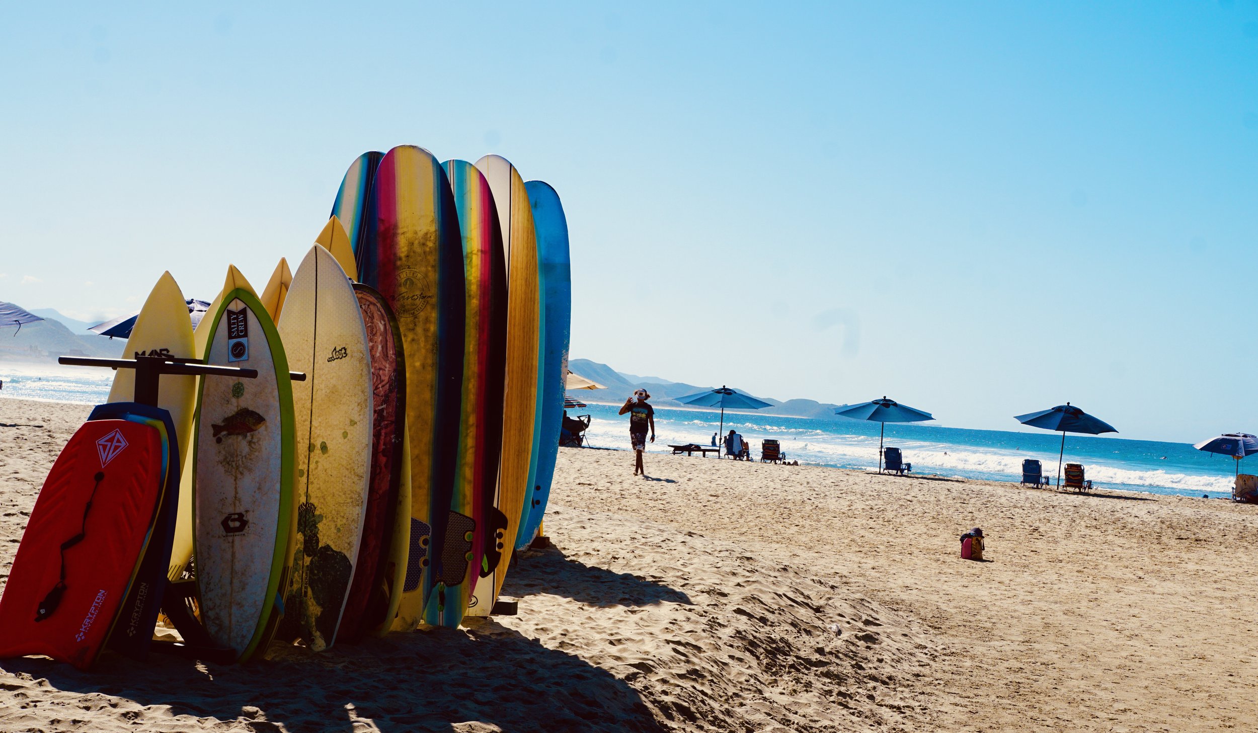 Colorful surfboards standing on the sandy beach near a clear blue sky, with people relaxing under umbrellas and a person walking along the shoreline.