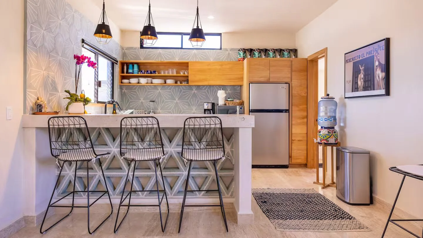 Modern kitchen with a marble breakfast bar, three black wire chairs, wooden cabinets, stainless steel refrigerator, water dispenser, and a wall-mounted TV.