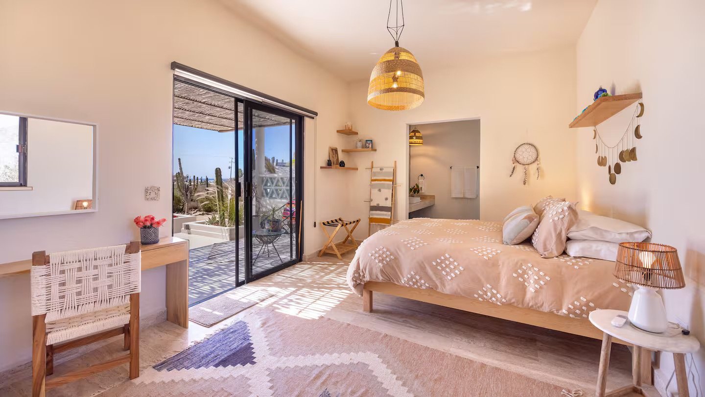 A cozy bedroom with a bed, bedside table with lamp, and a sliding glass door leading to a patio with outdoor furniture and cactus plants.