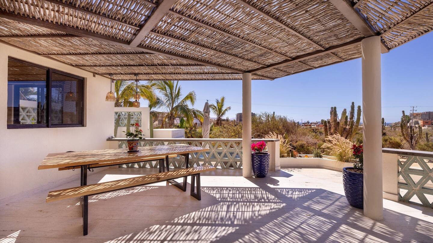 A covered outdoor patio with a wooden picnic table and benches, potted plants, and a view of palm trees, cacti, and a clear blue sky.