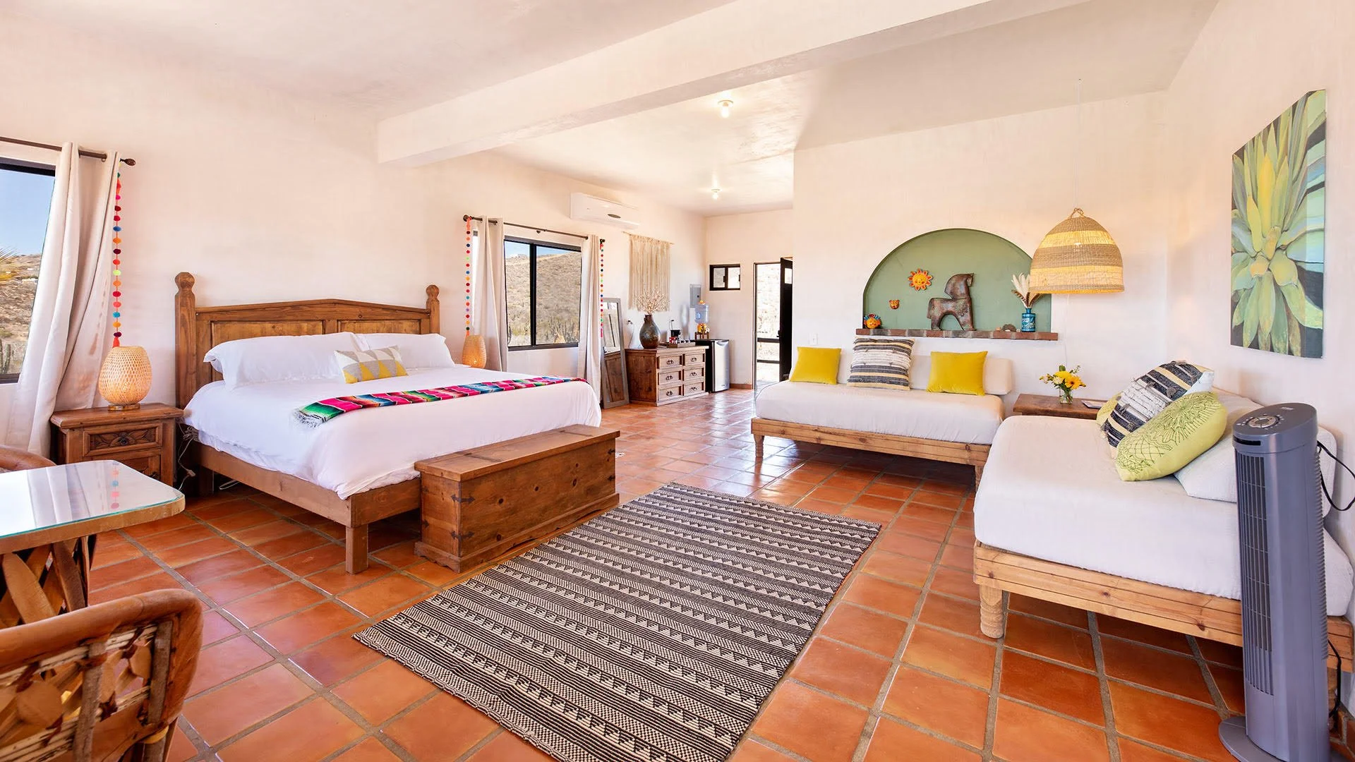 A spacious living area with a large bed, white bedding, and colorful pillows. There is a sitting area with a cushioned bench and pillows, a wooden side table with flowers, and decorative items on a shelf. The room has terracotta tile flooring, large 