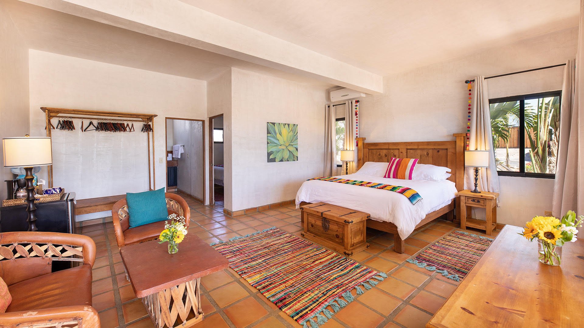 A cozy bedroom featuring a wooden bed with colorful striped blanket and pillows, two nightstands with lamps, a window with curtains showing palm trees outside, a small wooden chest at the foot of the bed, and decorative rugs on tiled floor. There's a