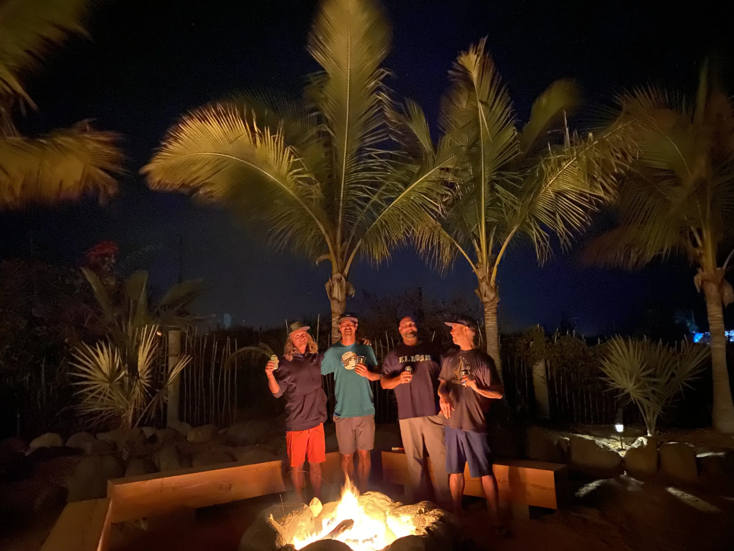 Group of people gathered around a fire pit outdoors at night, surrounded by palm trees, with some holding drinks and chatting.