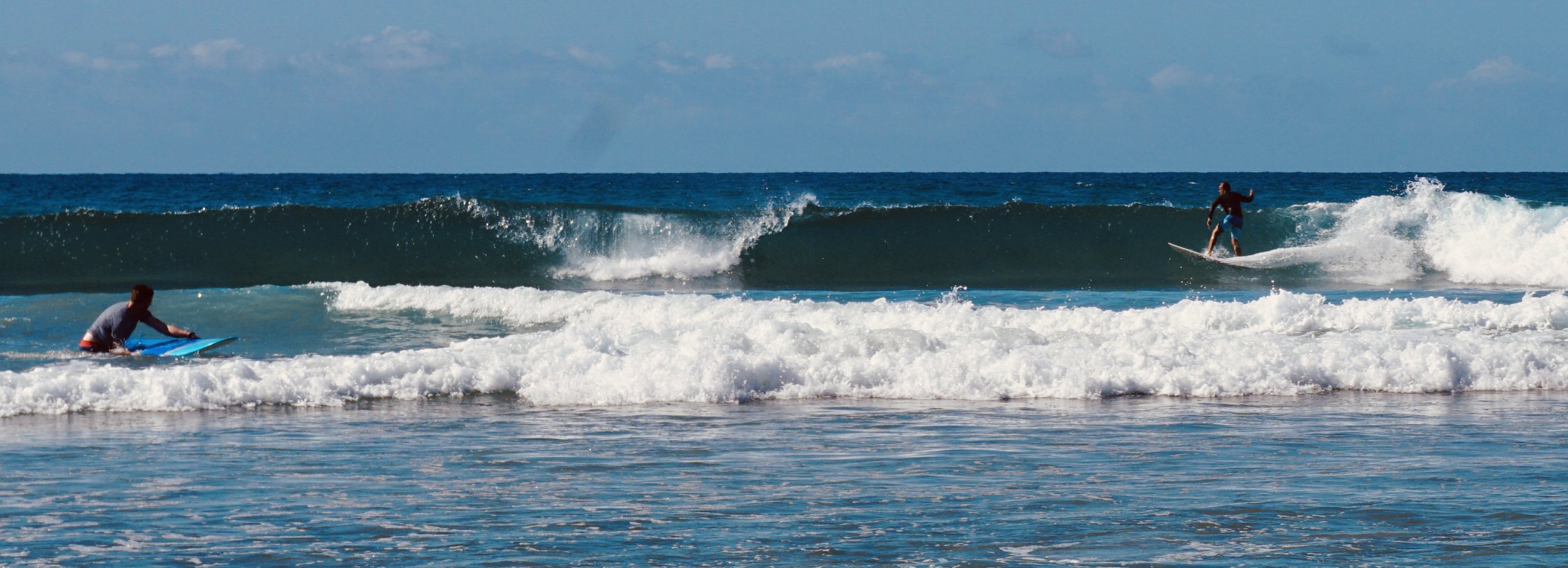 Two people surfing on ocean waves with one in the water on a surfboard and another riding a wave on a surfboard in the background.