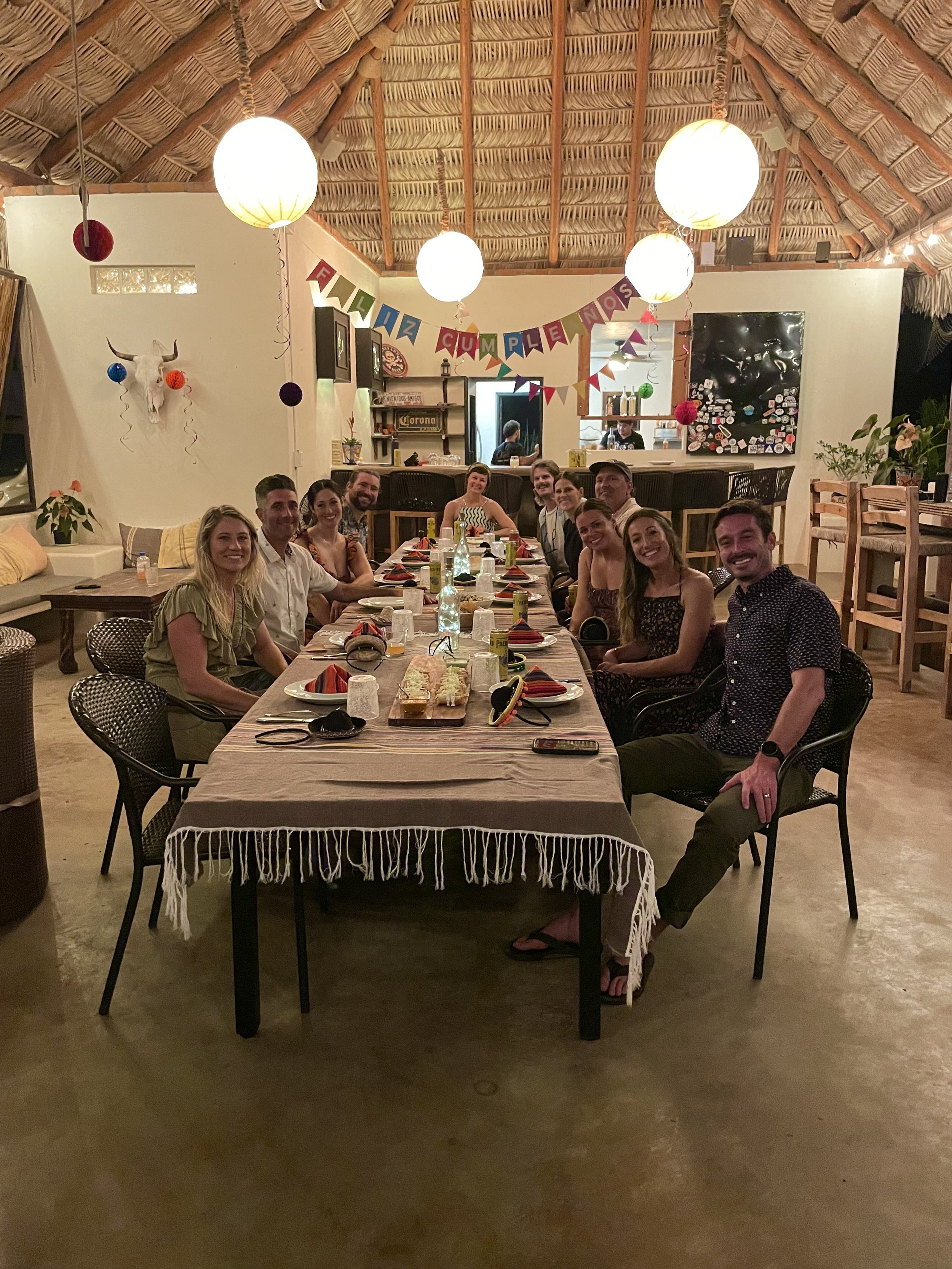 People gathered around a dining table decorated with a 'Vince Cumpleaños' banner, celebrating a birthday in a room with a thatched roof, hanging lanterns, and festive decorations.