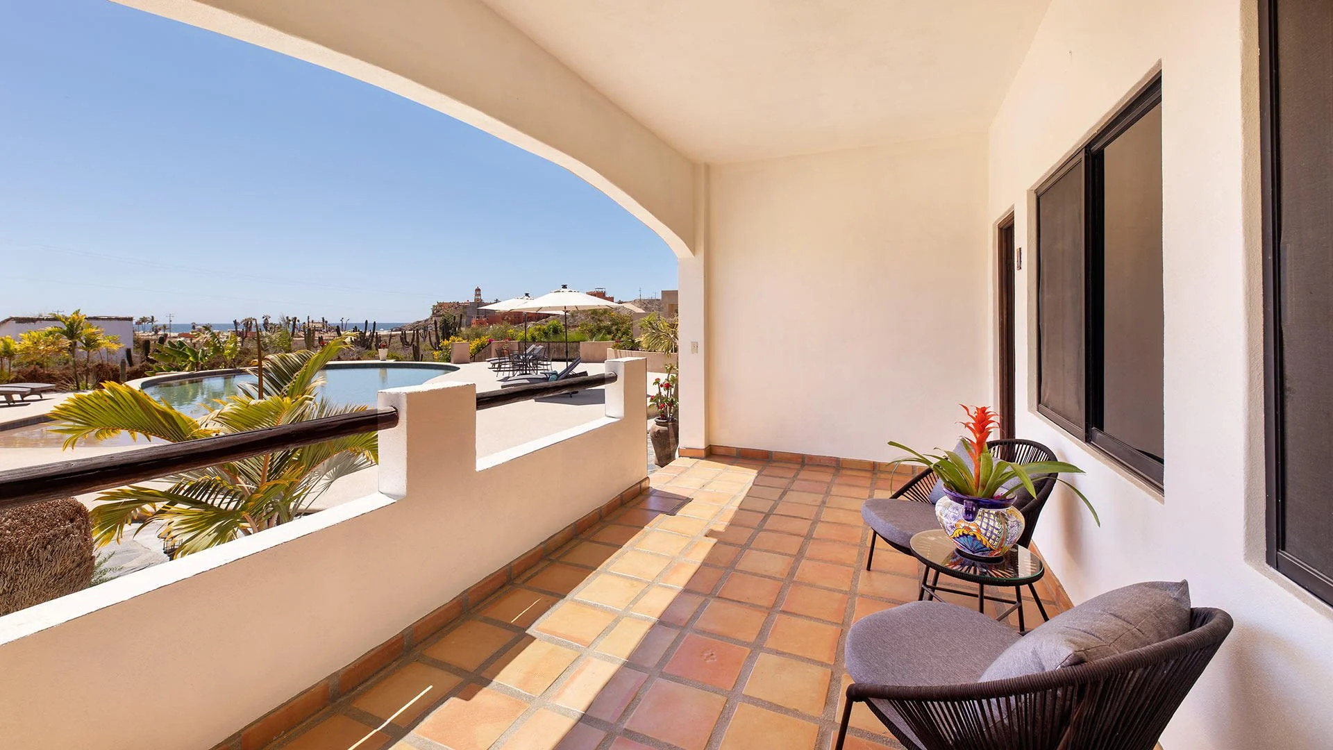Covered balcony with terracotta tiles, outdoor chairs, potted plants, view of a pool, palm trees, and a lighthouse in the distance under a clear blue sky.