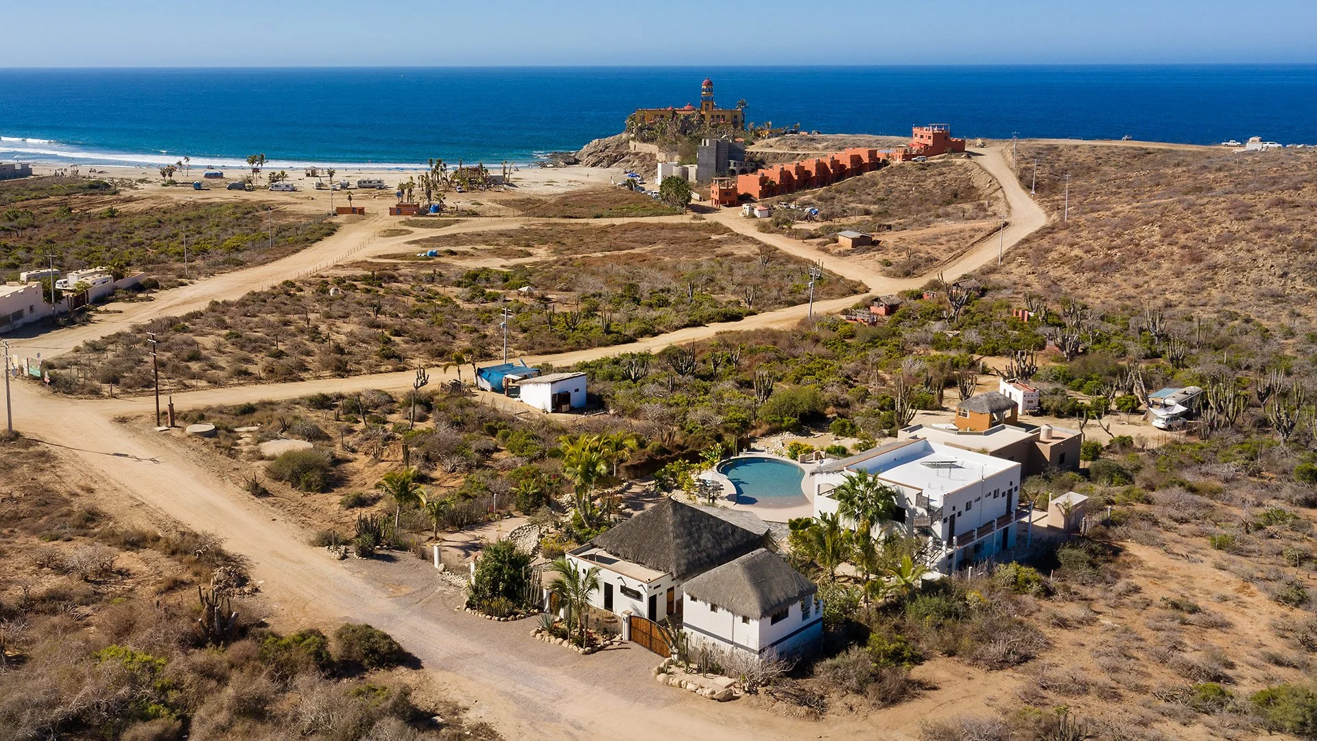 Aerial view of a beachside area with dirt roads, residential houses, and a large lighthouse on a cliff, overlooking the ocean with waves.