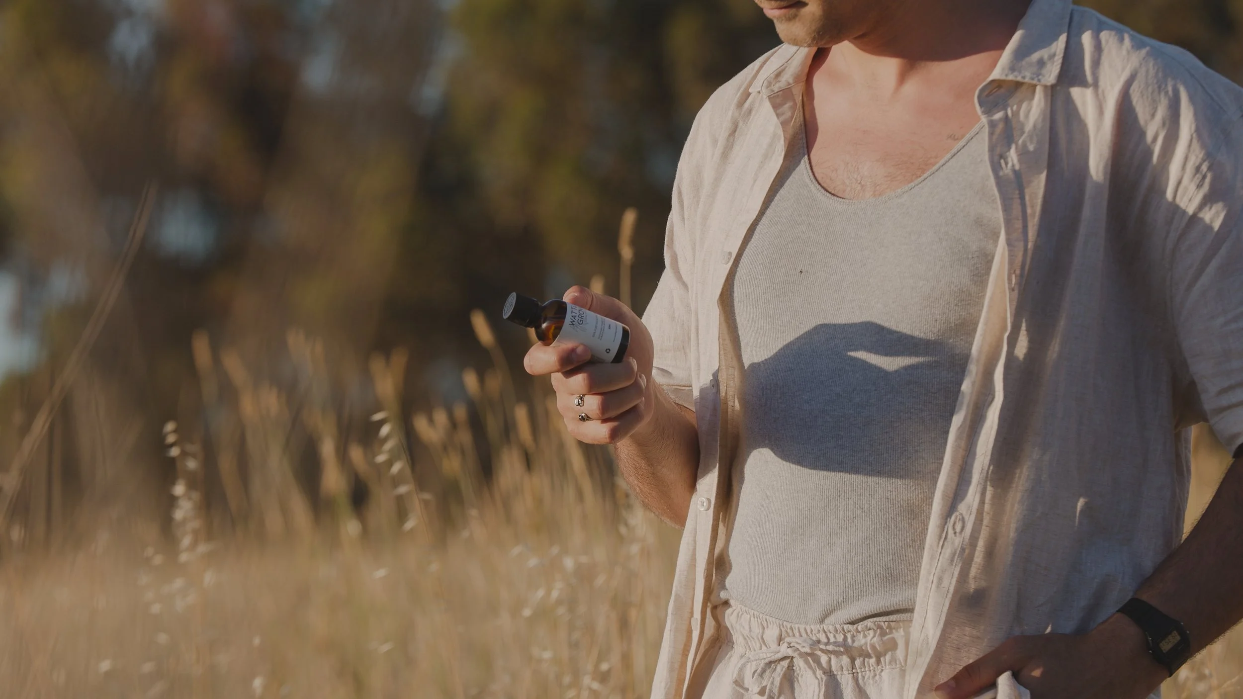 A man in a casual outfit holding a eucalyptus oil bottle in a field of tall grass during daylight.