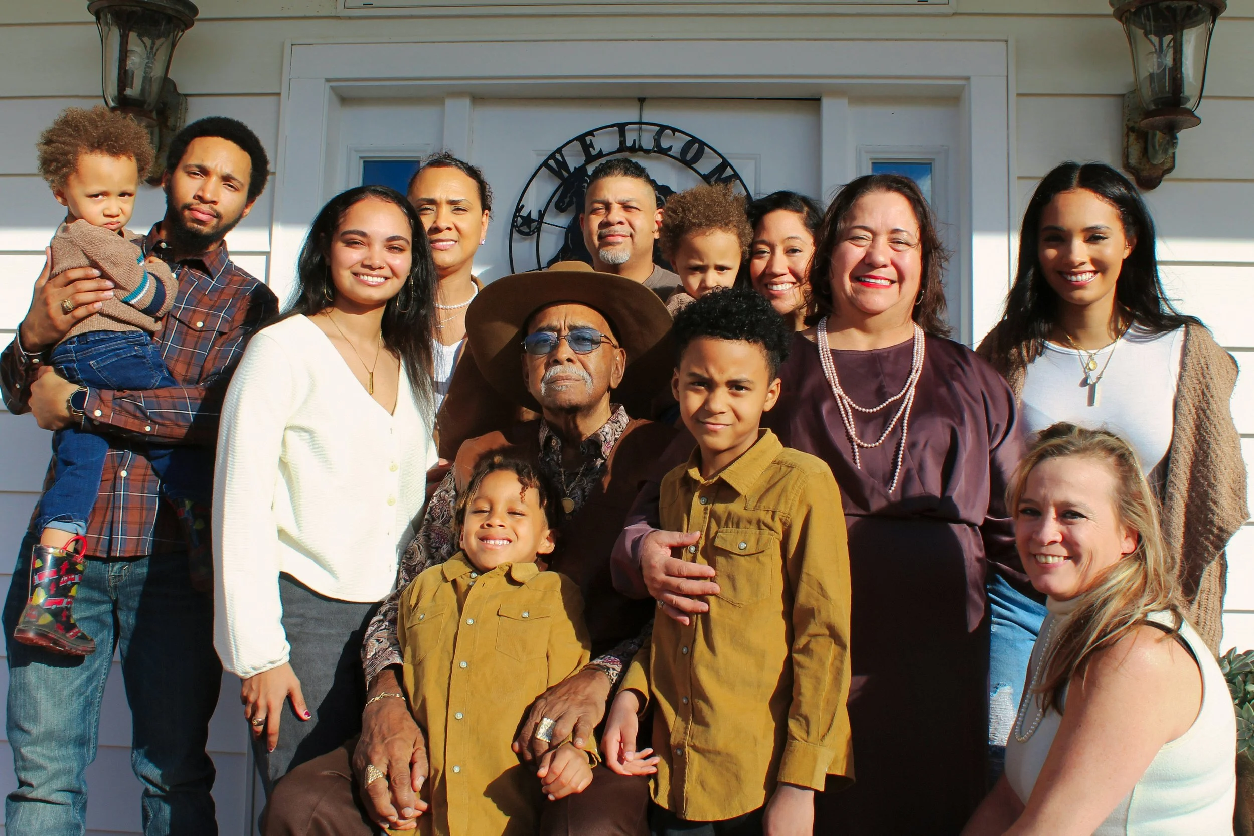 Multigenerational family group portrait outside a home, with an older man seated in front and adults and children gathered around him in bright sunlight.
