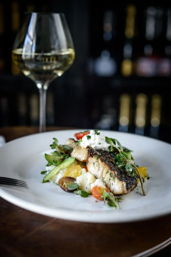 A plated dish featuring a cooked fish fillet, garnished with herbs, vegetables, and dollops of cream, with a glass of white wine in the background.
