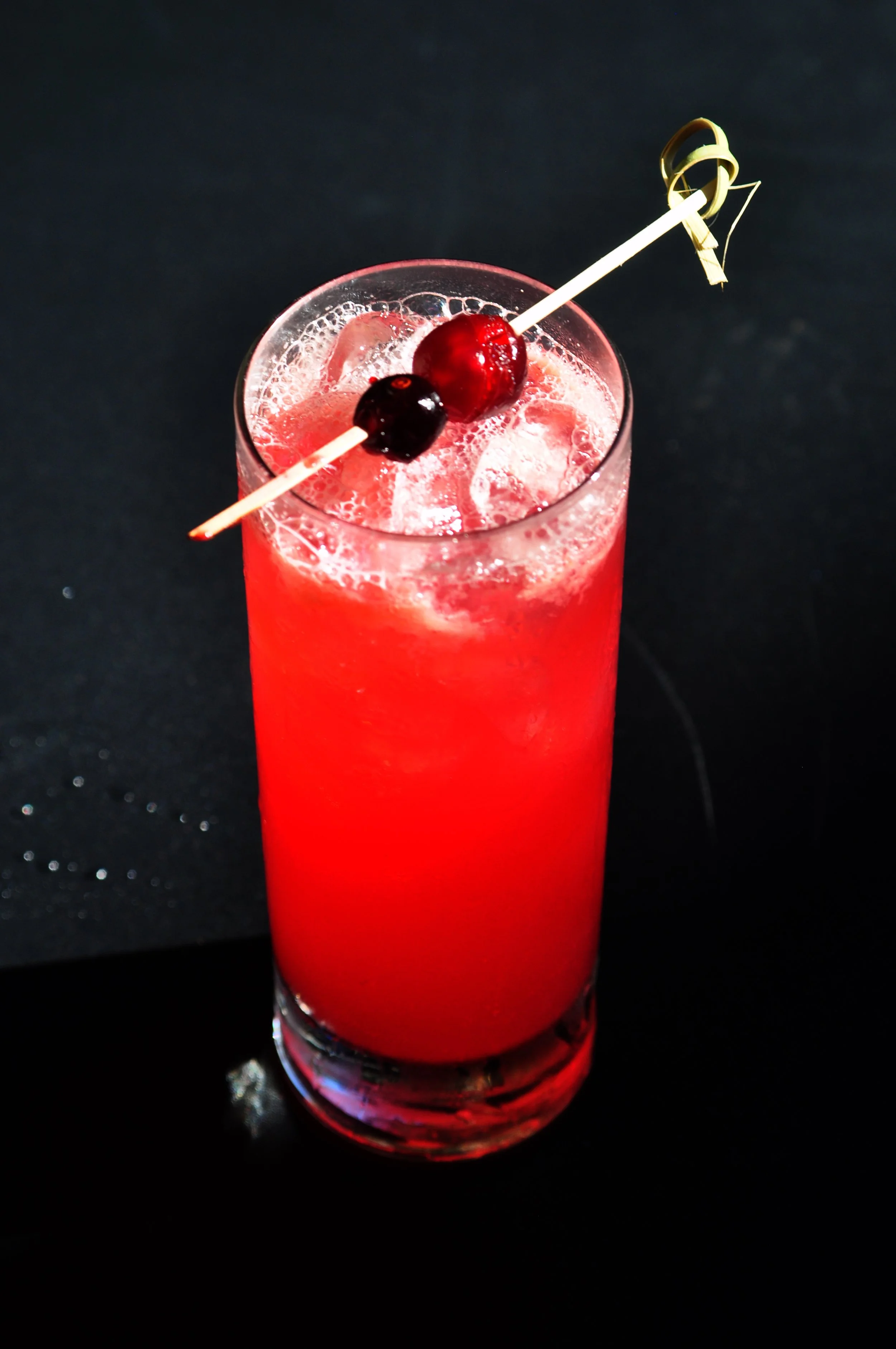 Top view of a Red colored cocktail in a high ball glass with skewed cherries on a black ground