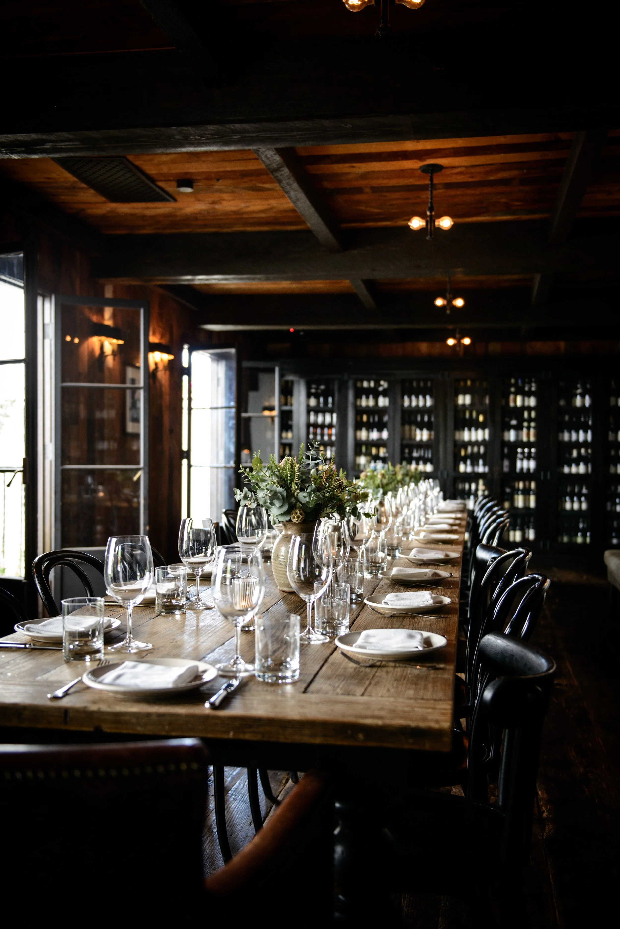 Elegant dining setup with a long wooden table, wine glasses, white napkins, and a floral centerpiece in a dimly lit room with wine racks and windows.