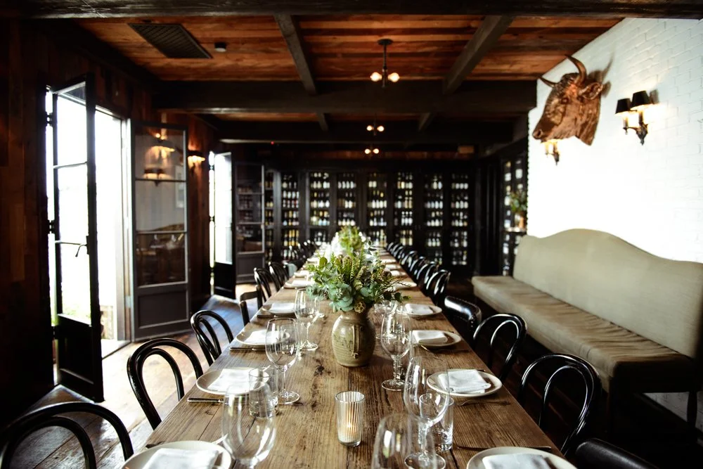 Long wooden dining table set with wine glasses, white plates, and a flower centerpiece in a rustic restaurant interior with wooden ceiling and white brick wall with mounted deer head.