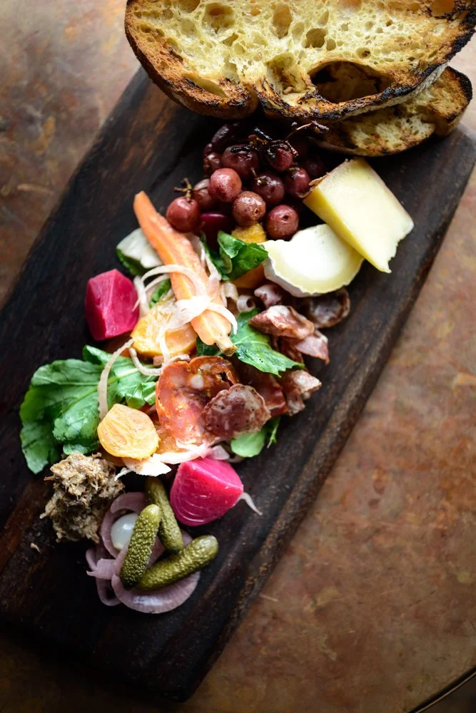 Assorted cheeses, pickles, cured meats, grapes, bread, and vegetables on a wooden serving board
