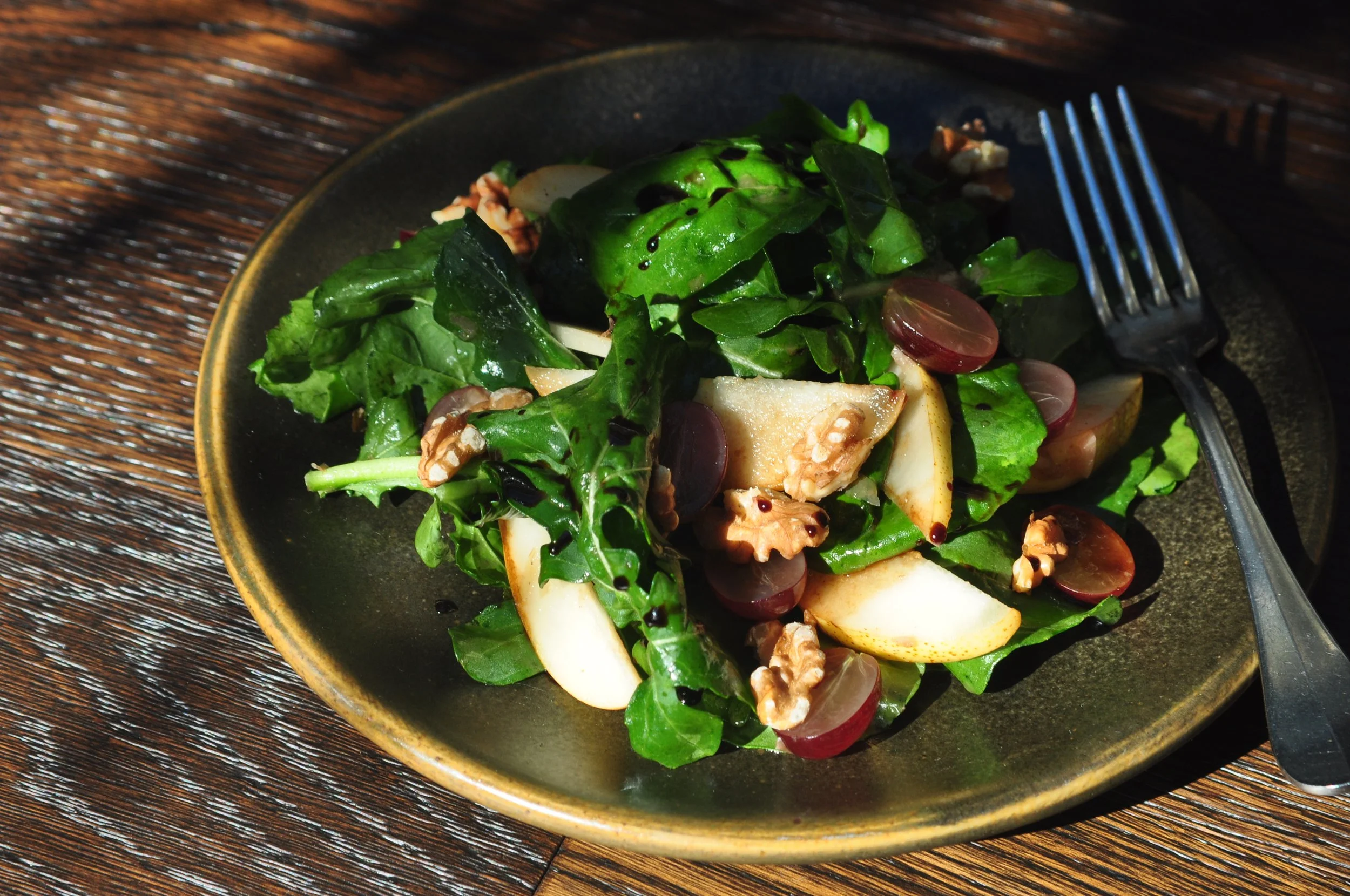 arugula, becky’s pears, grapes, walnuts & date syrup served in an olive green plate with fork on a rustic wood table background