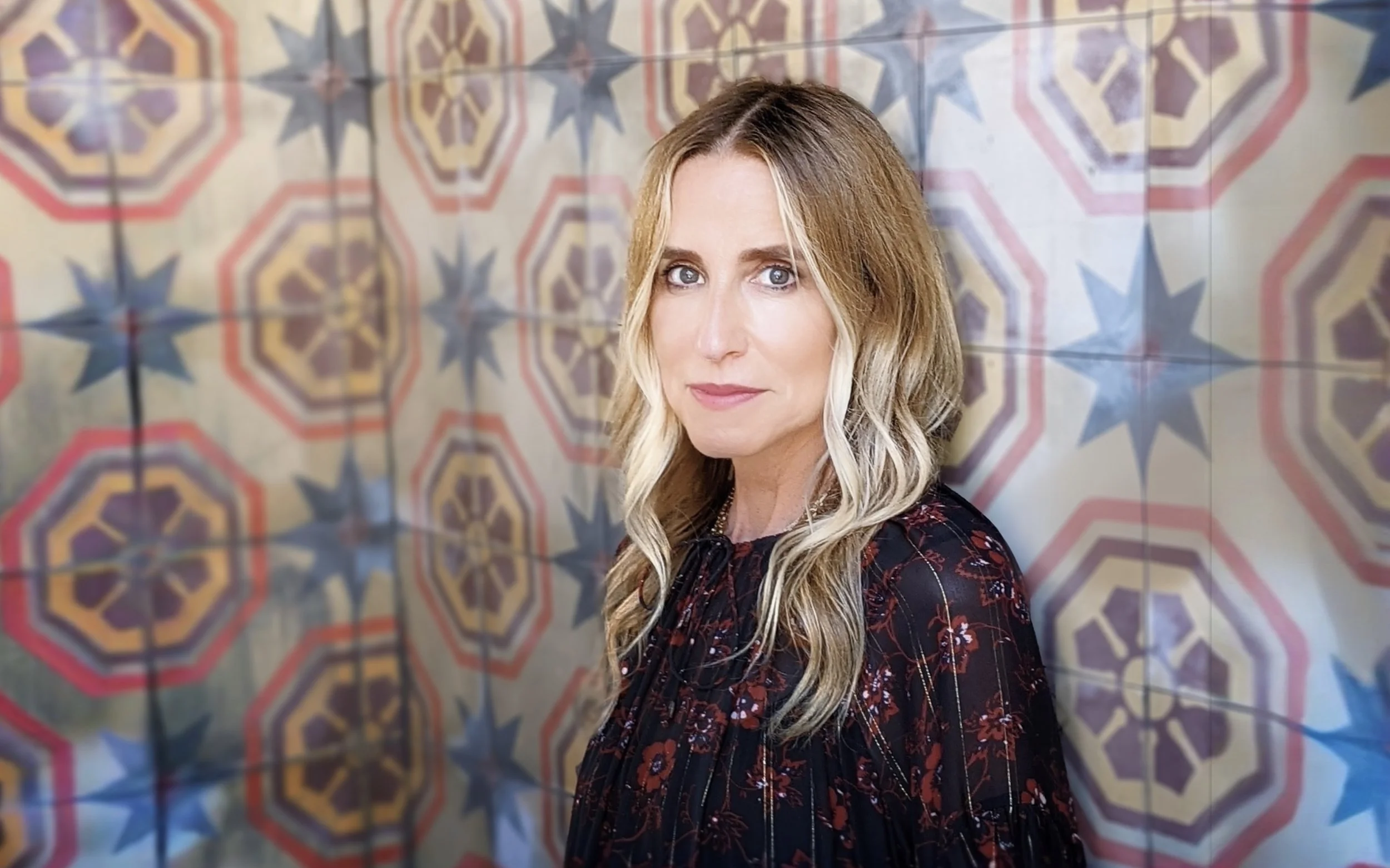 James Beard award winning restaurateur Caroline Styne, wearing a dark floral top, stands in front of a colorful tiled wall with geometric patterns.