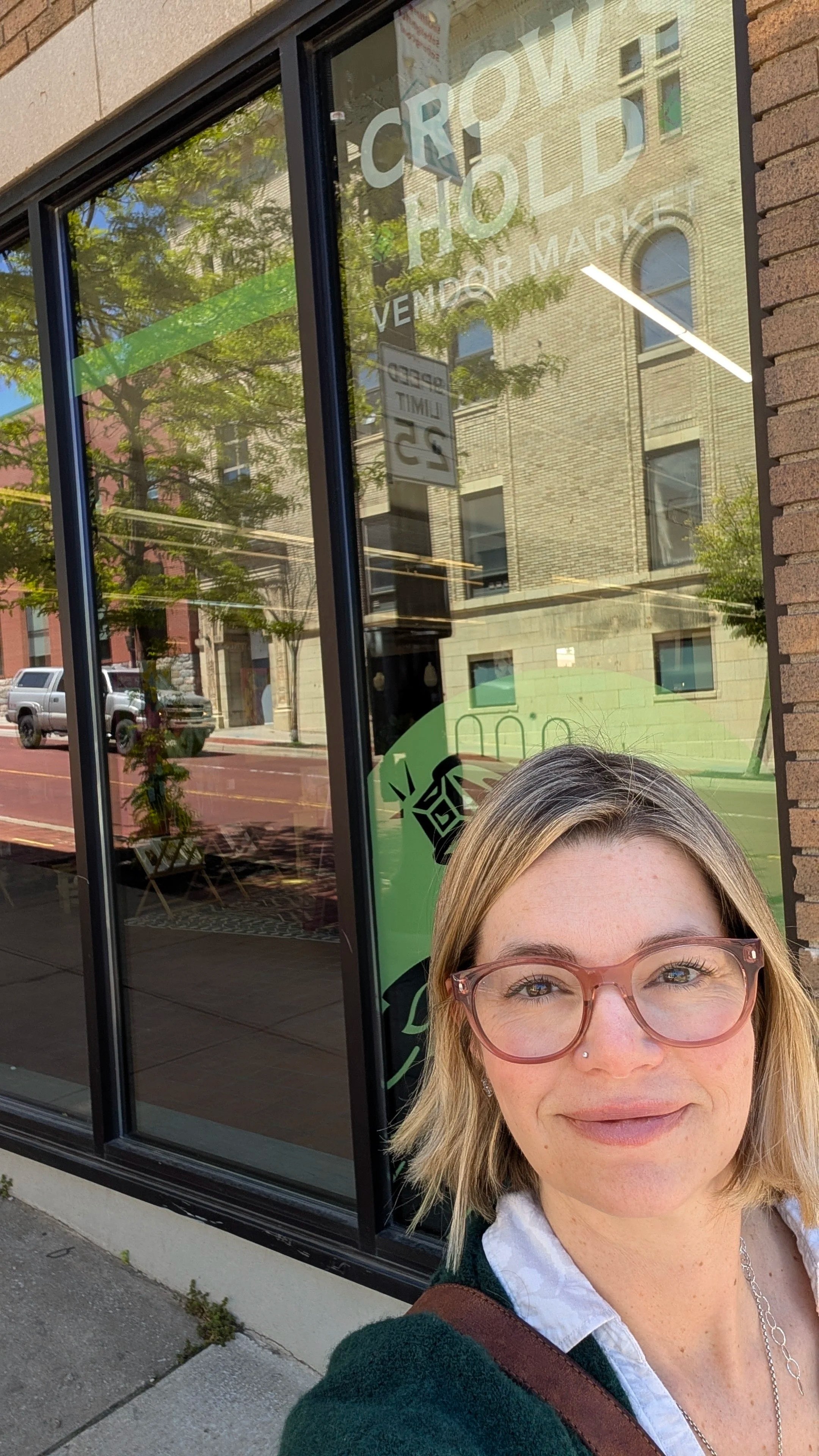 A woman with blonde hair, glasses, and a nose piercing taking a selfie in front of a store window with a "Crown Hold" sign, reflecting buildings, trees, and vehicles on the street.