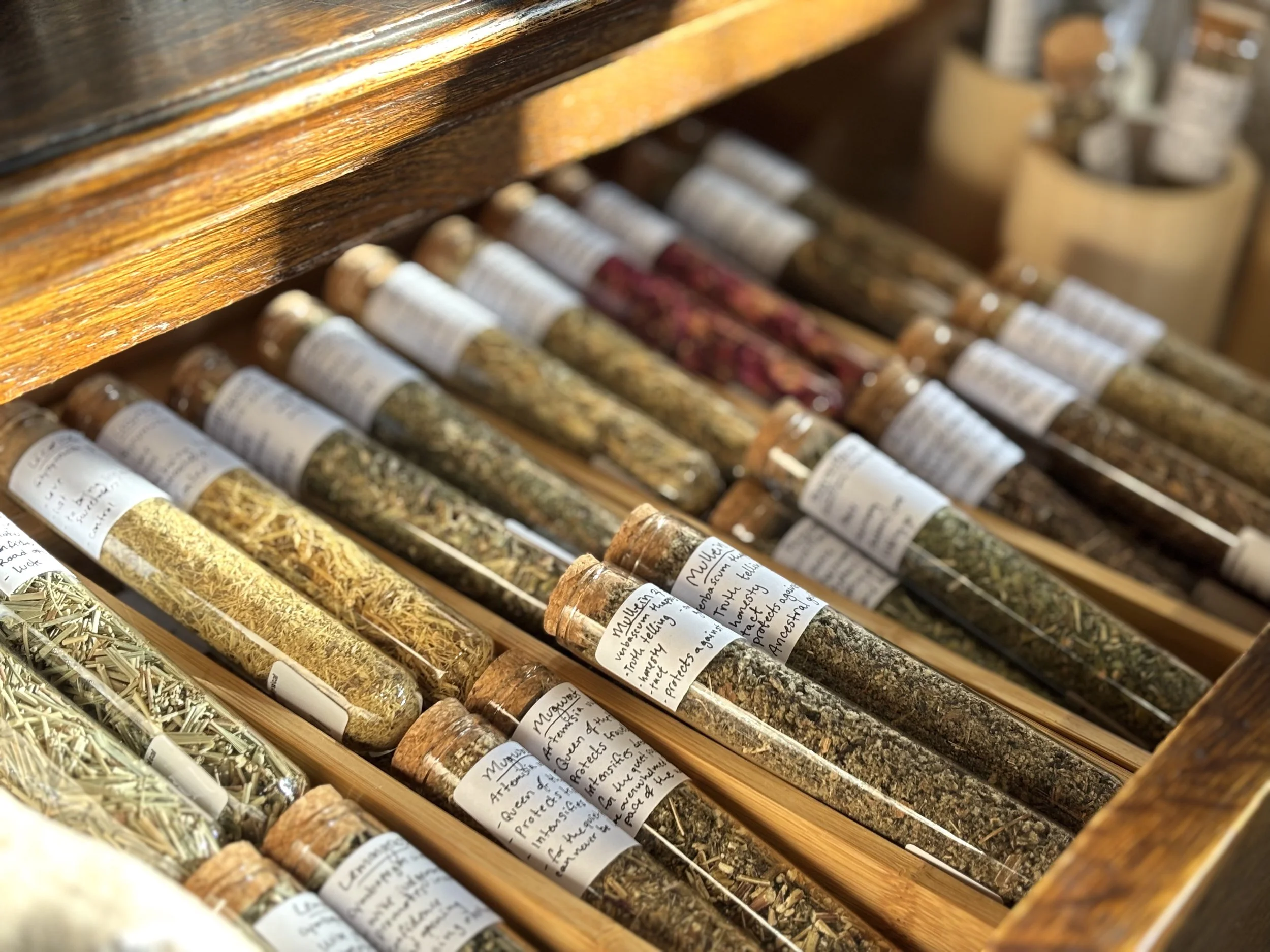 Collection of glass test tubes filled with various dried herbs and labeled with handwritten descriptions, stored in a wooden box.
