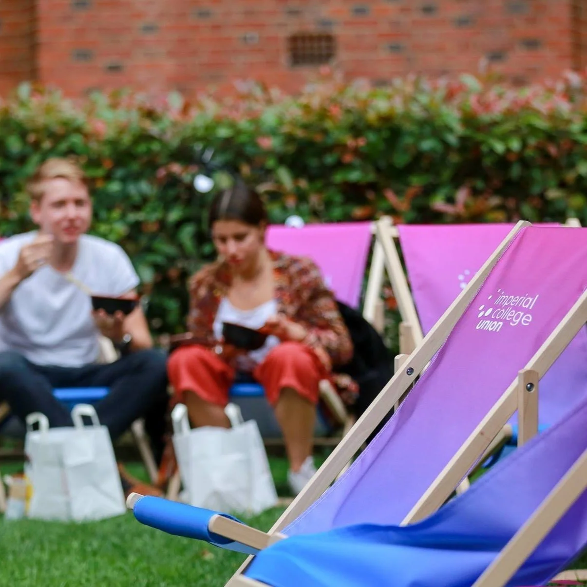 Pink and purple folding chairs with 'Imperial College Union' logo, two people sitting on a bench eating, green foliage, brick wall in background.