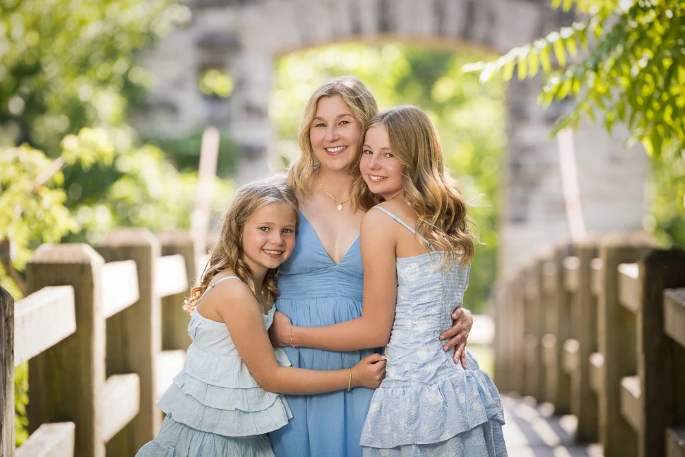 mom and two young daughters in blue sundresses hugging on a bridge