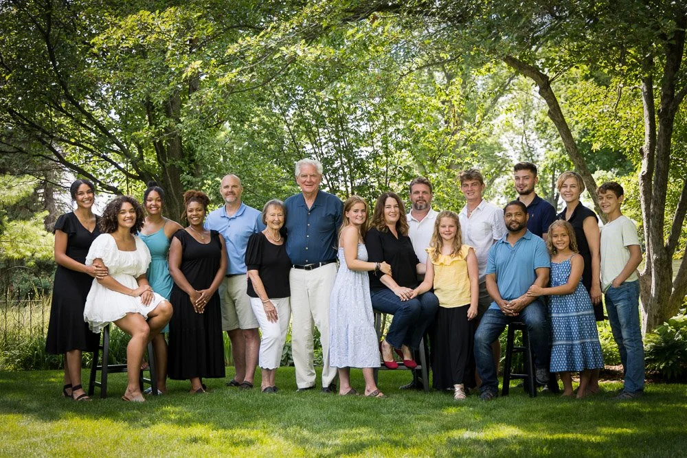 A large multigenerational family portrait outdoors under trees in a park, with people smiling and posing for the camera.