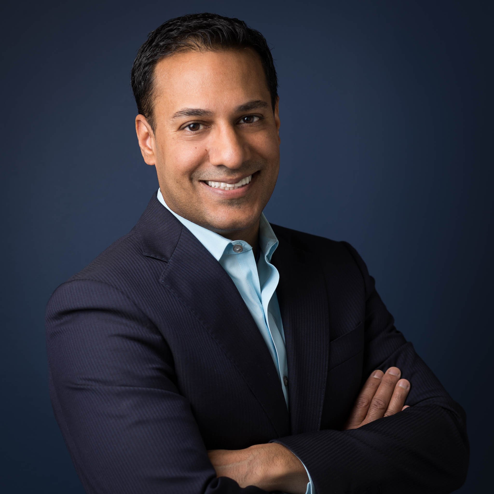 Man in blue suit with arms crossed smiles warmly in front of blue wall.