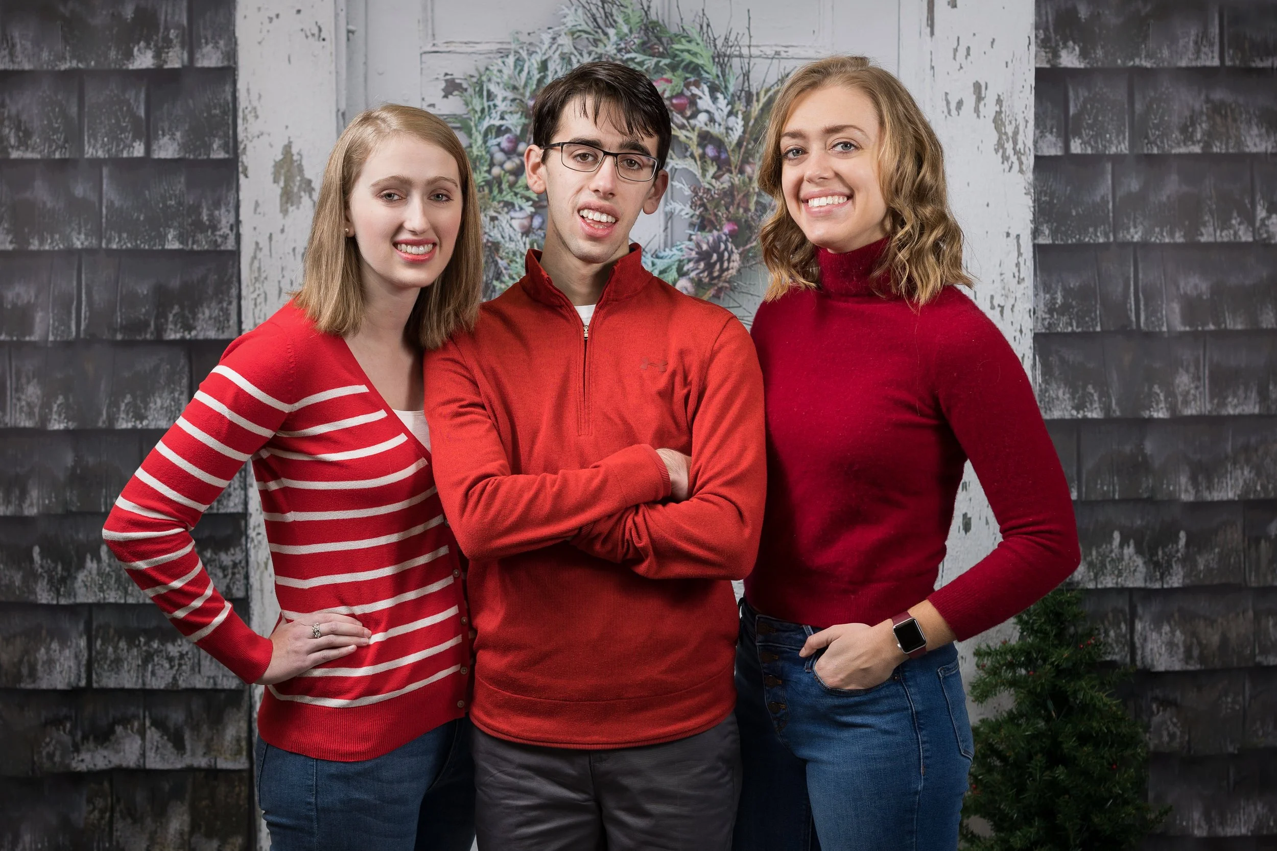 Three people smiling in front of a festive holiday backdrop with a wreath. The two women on either side are wearing red tops, and the man in the center is wearing a red sweater.