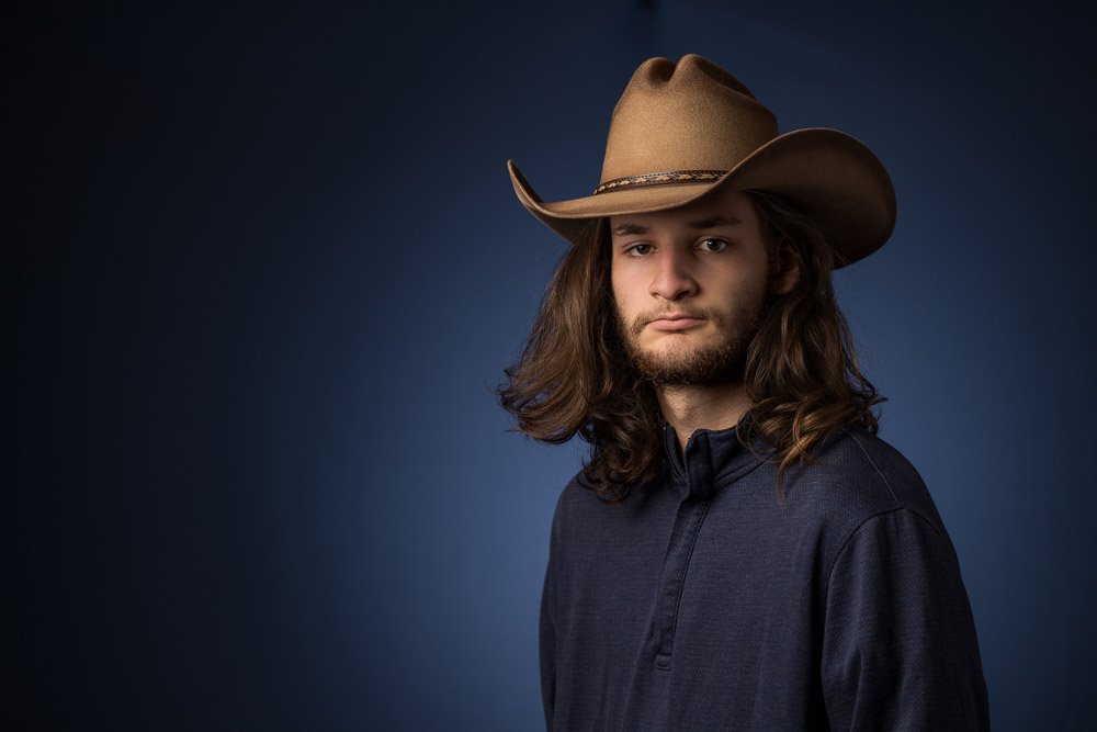 young man with long hair and a cowboy hat in front of a blue wall