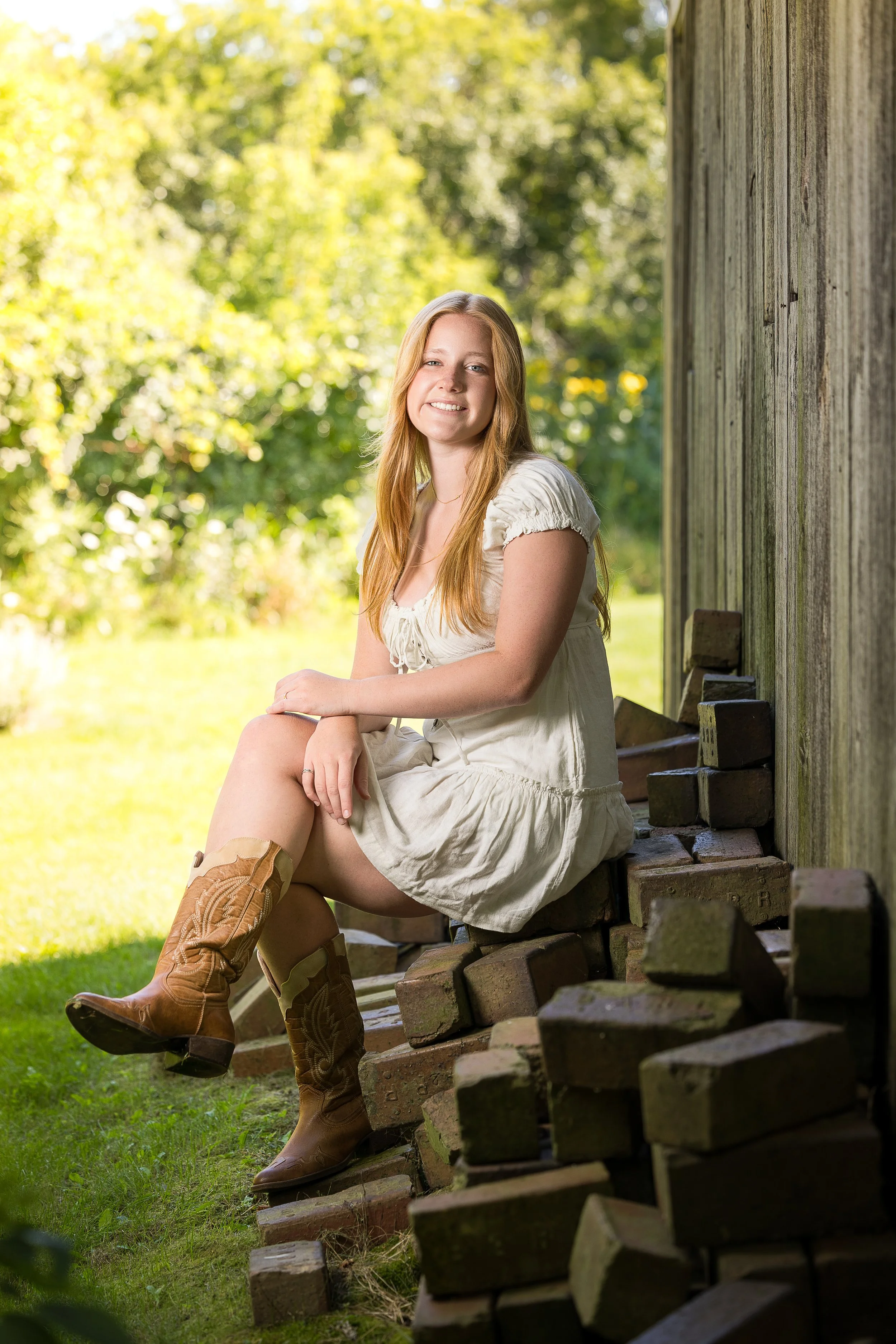 A woman in a white dress and cowboy boots sitting on bricks next to a wooden wall, with a green, sunny garden in the background.
