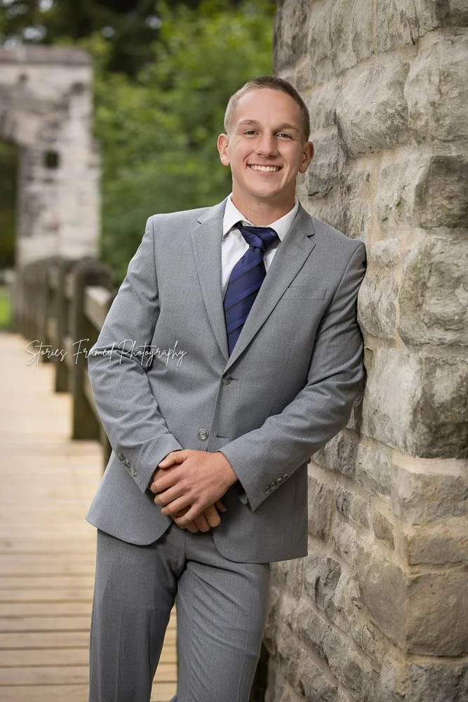 Wauwatosa West senior boy in a gray suit and navy tie leaning against a stone wall on a wooden bridge for his senior portraits near Milwaukee, Wisconsin