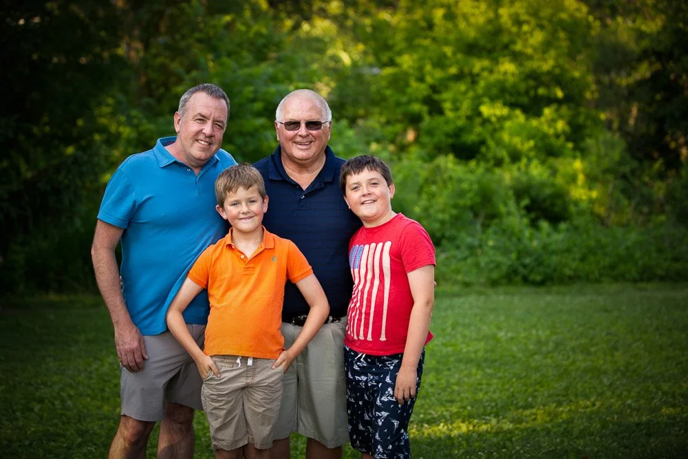 Four males, two adults and two children, standing outdoors on green grass with trees in the background, smiling for the photo.