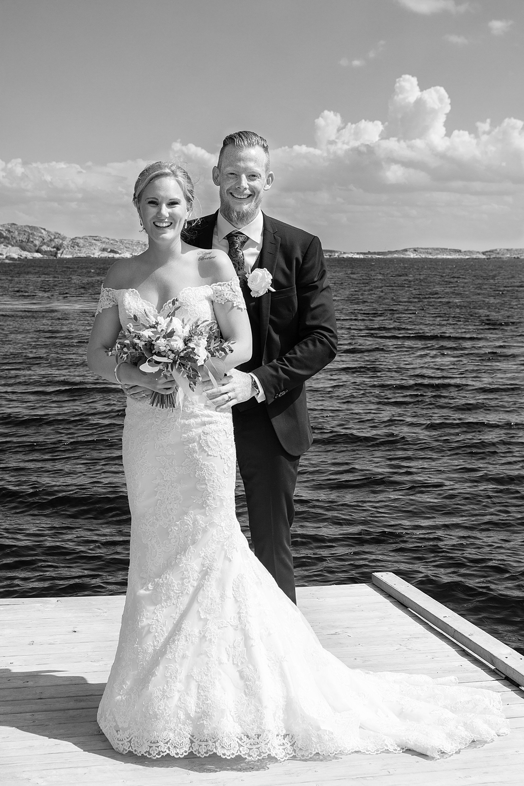 Bride and groom posing by the water on a sunny day, with the bride wearing a lace wedding dress and holding a bouquet, and the groom in a suit.