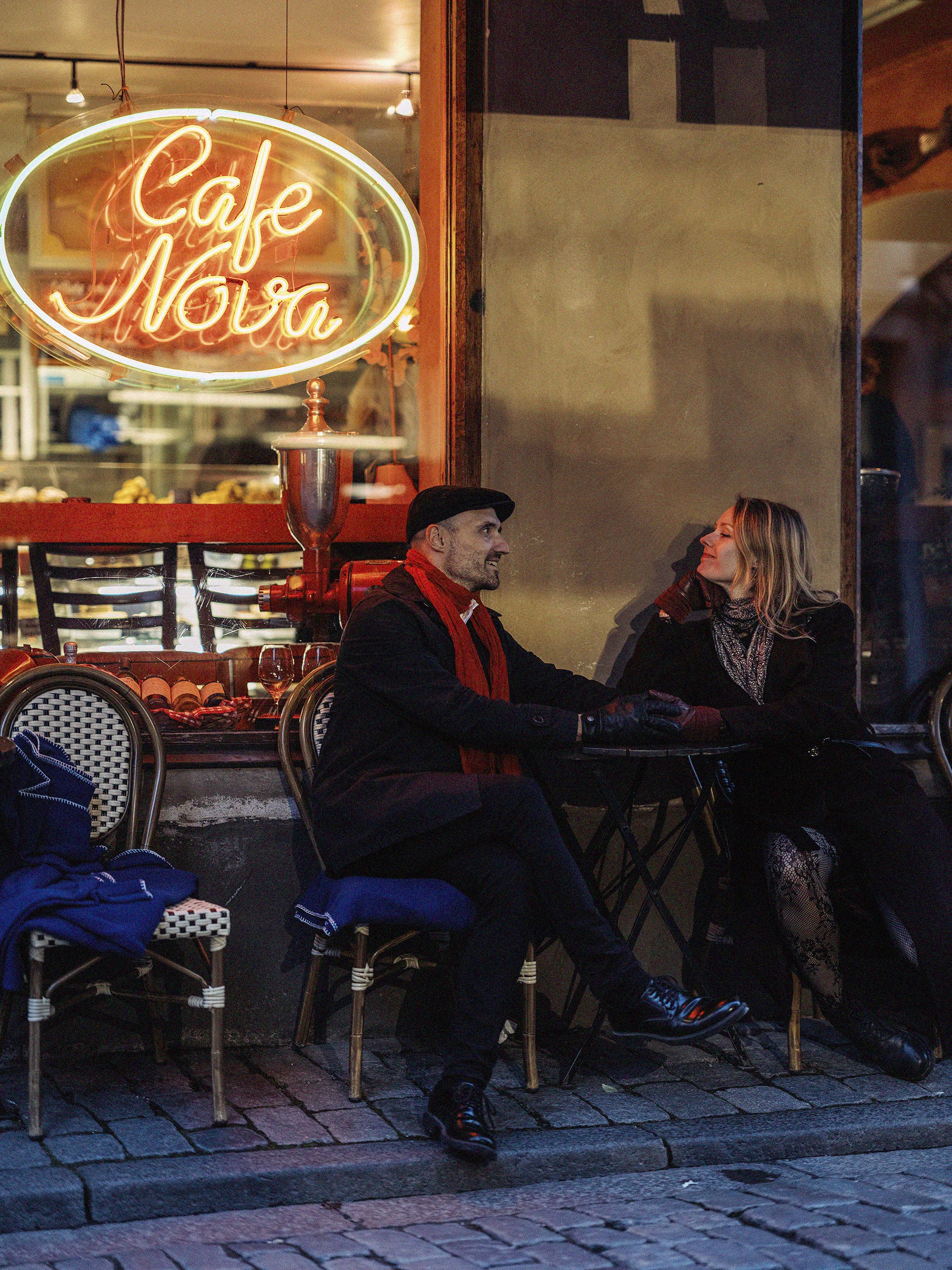 A man and woman sitting at an outdoor cafe table under a neon sign that reads "Cafe Nova." They appear to be engaged in conversation on a cobblestone street.