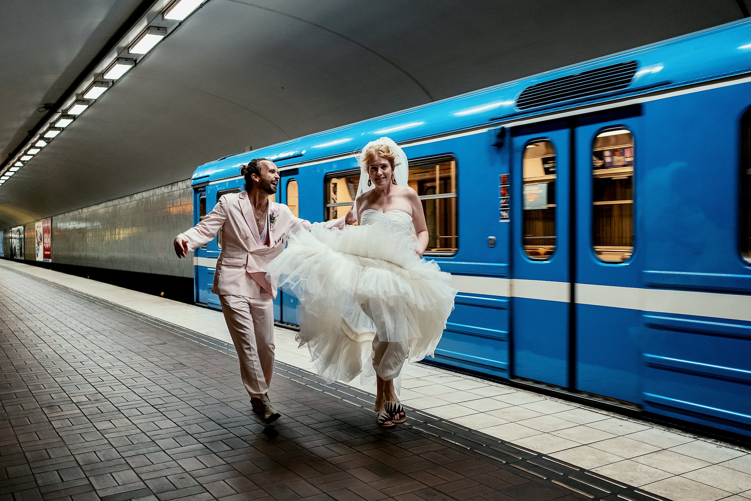 Bride and groom running in a subway station with a blue train in the background.